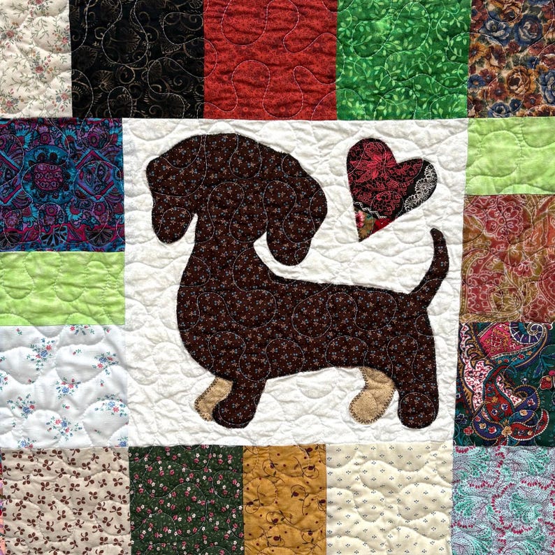 Pu&ograve; includere: Una trapunta patchwork con un'applicazione di bassotto marrone e un cuore. La trapunta &egrave; composta da vari quadrati di tessuto nei toni del rosso, verde, blu e bianco, con disegni floreali e fantasia. Il cane &egrave; lungo circa 25 cm.