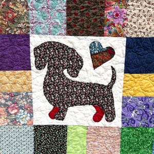 Pu&ograve; includere: Una trapunta patchwork con un'applicazione di bassotto in tessuto floreale nero e rosso. Un cuore di tessuto &egrave; posto accanto al cane. La trapunta &egrave; composta da quadrati di tessuto di diversi colori e motivi.