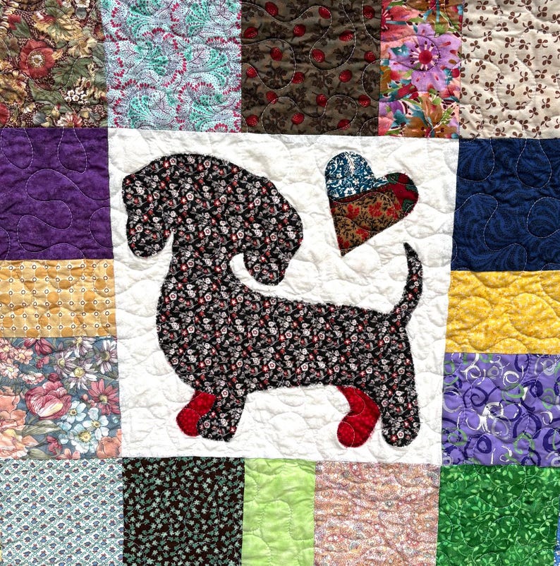 Pu&ograve; includere: Una trapunta patchwork con un'applicazione di bassotto in tessuto floreale nero e rosso. Un cuore di tessuto &egrave; posto accanto al cane. La trapunta &egrave; composta da quadrati di tessuto di diversi colori e motivi.