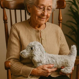 May include: A plush, gray dachshund dog toy with black eyes and a small green tag. The toy is held by a person wearing a tan cardigan and sitting in a wooden rocking chair. The background is a neutral color.