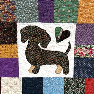 Pu&ograve; includere: Una trapunta patchwork con un motivo di bassotto. Il cane &egrave; realizzato in tessuto scuro con motivo floreale, su un quadrato bianco. La trapunta presenta quadrati con vari motivi nei toni del verde, viola e oro.