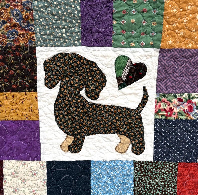 Pu&ograve; includere: Una trapunta patchwork con un motivo di bassotto. Il cane &egrave; realizzato in tessuto scuro con motivo floreale, su un quadrato bianco. La trapunta presenta quadrati con vari motivi nei toni del verde, viola e oro.