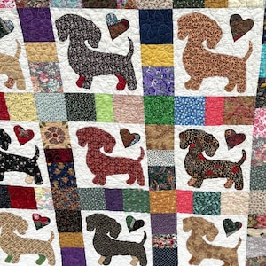 Pu&ograve; includere: Una trapunta patchwork con applicazioni di cani bassotto e cuori. I cani sono realizzati con vari tessuti fantasia, con ogni cane e cuore posti su un quadrato bianco. La trapunta ha un aspetto colorato e fatto a mano.