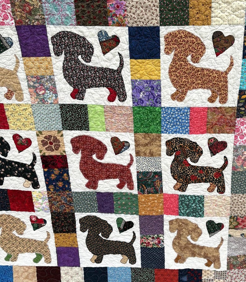 Pu&ograve; includere: Una trapunta patchwork con applicazioni di cani bassotto e cuori. I cani sono realizzati con vari tessuti fantasia, con ogni cane e cuore posti su un quadrato bianco. La trapunta ha un aspetto colorato e fatto a mano.