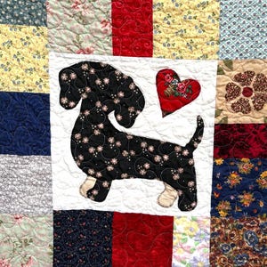 Pu&ograve; includere: Una trapunta patchwork con un disegno di bassotto nero e un cuore rosso. La trapunta &egrave; composta da quadrati di tessuto fantasia, tra cui motivi floreali e colori uniti, con cuciture trapuntate. Il bassotto ha accenti floreali.