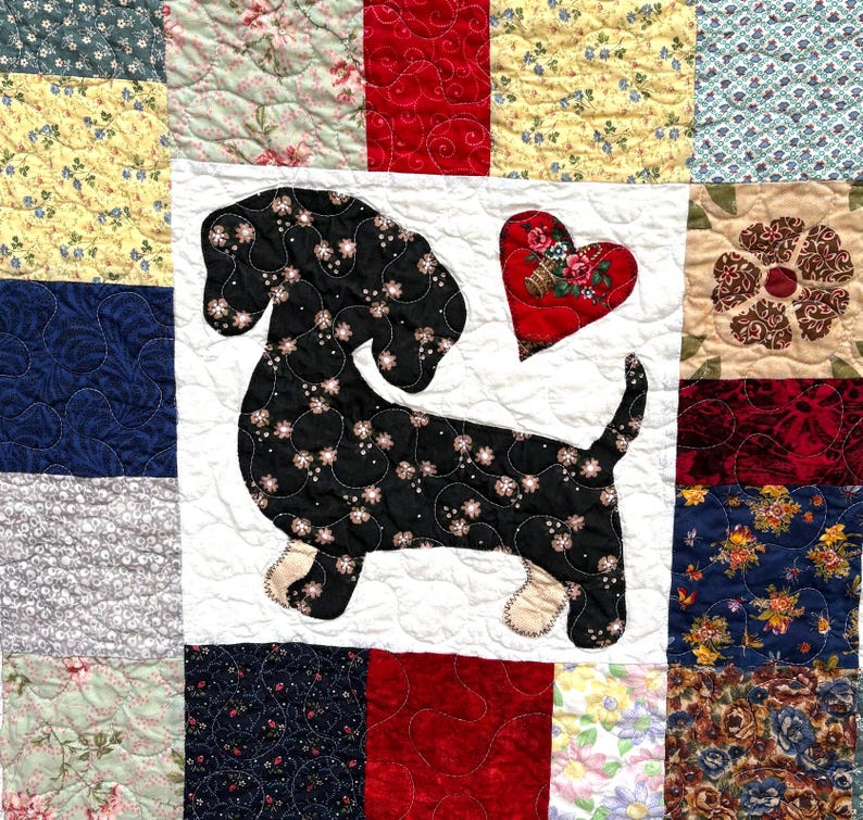 Pu&ograve; includere: Una trapunta patchwork con un disegno di bassotto nero e un cuore rosso. La trapunta &egrave; composta da quadrati di tessuto fantasia, tra cui motivi floreali e colori uniti, con cuciture trapuntate. Il bassotto ha accenti floreali.