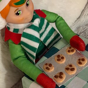 May include: A Christmas elf doll wearing a green and white striped scarf, green outfit, and red shoes. The elf is holding a baking sheet with six chocolate chip cookies on a green and white checkered surface.