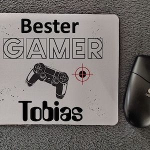 Könnte beinhalten: Ein weißes Mauspad mit schwarzem Text, der "Bester GAMER Tobias" lautet, und einer Schwarzweißillustration eines Videospielcontrollers und einer Zielscheibe.