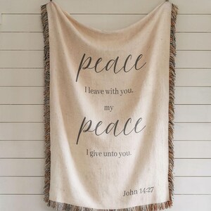 Könnte beinhalten: Cremefarbene gewebte Decke mit dem Wort „Frieden“ in Schreibschrift. Die Decke hat einen Fransenrand und enthält den Text „Ich lasse euch Frieden, meinen Frieden gebe ich euch. Johannes 14,27."