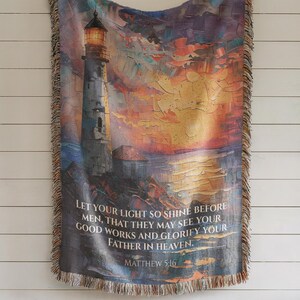 May include: Woven throw blanket depicting a lighthouse against a sunset. The blanket has a fringed edge and includes the text "LET YOUR LIGHT SO SHINE BEFORE MEN, THAT THEY MAY SEE YOUR GOOD WORKS AND GLORIFY YOUR FATHER IN HEAVEN. MATTHEW 5:16."