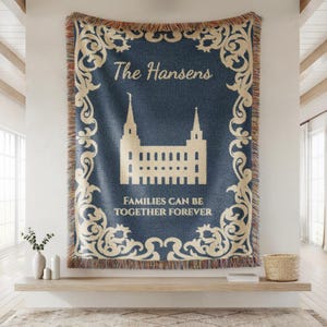 May include: A navy blue woven throw blanket with a cream-colored decorative border and text. The blanket features a cream-colored illustration of a building with two spires and the words "The Hansens" and "FAMILIES CAN BE TOGETHER FOREVER".