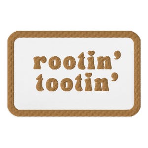 Könnte beinhalten: Gestickter Aufnäher mit den Worten "rootin' tootin'" in braunem Faden auf weißem Hintergrund. Der Aufnäher hat einen braunen Rand. Der Text ist in einer verspielten, abgerundeten Schriftart.