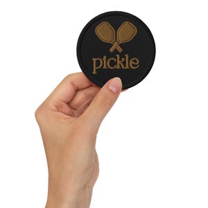 May include: A black circular patch with a brown embroidered pickleball paddle design and the word "pickle" in brown embroidery.