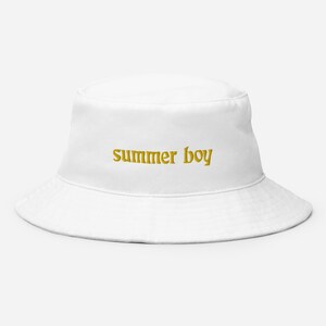 Puede incluir: Un gorro de pescador blanco con el texto "summer boy" bordado en dorado en la parte delantera.