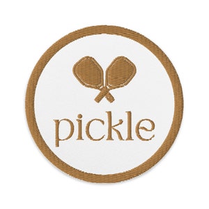 Könnte beinhalten: Ein brauner gestickter Aufnäher mit weißem Hintergrund. Der Aufnäher zeigt zwei gekreuzte Pickleball-Schläger und das Wort "pickle" in brauner Stickerei.