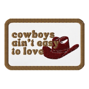 May include: Embroidered patch with a white background and a brown border. The patch features the text "cowboys ain't easy to love" in brown letters, along with a red cowboy hat.