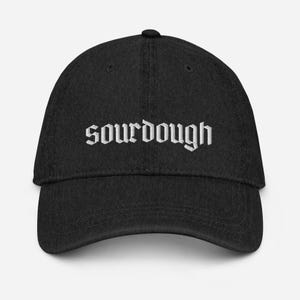 Può includere: Cappellino da baseball nero con la parola "sourdough" ricamata in bianco con lettere gotiche sul davanti. Il cappellino ha una visiera curva e una corona arrotondata.