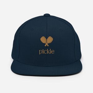 Puede incluir: Una gorra de béisbol azul marino con un diseño de pala de pickleball bordado en marrón y la palabra "pickle" en marrón.