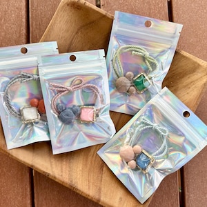 May include: Four hair scrunchies in iridescent plastic bags, each with a different design. The scrunchies have decorative elements like square gems and small, round beads. The colors include pink, blue, green, and gray.