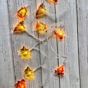 Puede incluir: Cadena de luces de hadas con temática de camping. Las luces presentan imágenes de una autocaravana, una tienda de campaña, una fogata y una linterna. Las luces están enhebradas en un cable blanco y están iluminadas con luces LED blanco cálido.