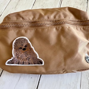 May include: A tan fanny pack with a brown zipper and a Chewbacca patch. The patch is an embroidered image of Chewbacca. The fanny pack has a tan strap and a silver-colored metal emblem.