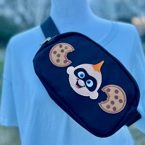 Può includere: Marsupio nero con un personaggio dei cartoni animati di un bambino che indossa una maschera e tiene dei biscotti. Il personaggio è circondato da due biscotti con morsi.