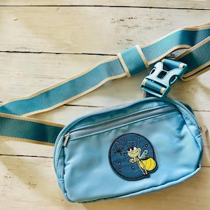 May include: Light blue fanny pack with a matching adjustable strap and buckle. The pack features a circular patch with a cartoon firefly design. The strap has a light beige stripe down the center.