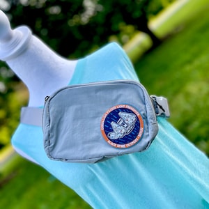 Puede incluir: Una riñonera gris con un parche negro y azul que dice "Corellian YT-1300 Light Freighter Black Spine Outpost - Batuu".