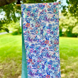 Könnte beinhalten: Ein gemusterter Stoff mit floralem und mechanischem Design hängt an einem Holzbügel. Der Stoff hat eine weiße obere Schicht mit einer türkisfarbenen Rückseite. Das Design umfasst Blumen, Zahnräder und mechanische Elemente in Blau, Rosa und Türkis.