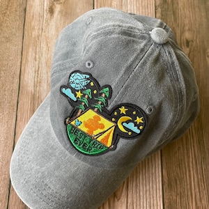 Könnte beinhalten: Eine graue Baseballkappe mit einem gestickten Aufnäher einer Mickey-Maus-Kopf mit einem Zelt, Sternen und dem Text "Best Trip Ever".