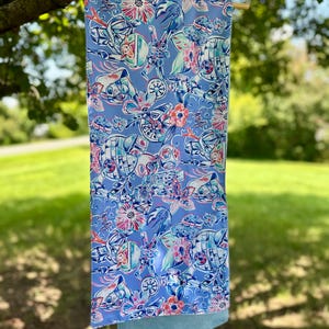 Könnte beinhalten: Ein hellblaues Dekohandtuch mit einem farbenfrohen Muster mit floralen und Star-Wars-Illustrationen. Das Handtuch hängt an einer Holzstange, mit einem hellblauen unteren Abschnitt. Der Hintergrund ist ein verschwommener grüner Rasen und Bäume.