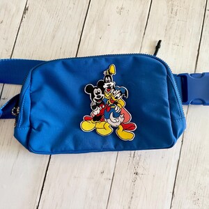 Peut inclure: Sac banane bleu vif avec une sangle réglable et une boucle. Il présente une application colorée de Mickey Mouse, Dingo et Donald Duck. Le sac est fait d'un matériau durable et résistant à l'eau.