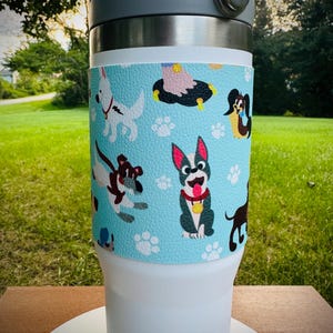 May include: A white tumbler with a light blue wrap featuring cartoon dogs and paw prints. The tumbler has a grey lid and handle. The background is a grassy area.