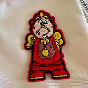 Peut inclure: Écusson brodé représentant un personnage de dessin animé en tenue rouge avec des détails jaunes. Le personnage a un visage d'horloge et une expression souriante. L'écusson est sur un fond beige.