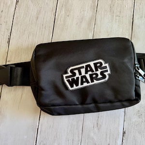 Puede incluir: Riñonera negra con un parche con el logo de Star Wars. La riñonera tiene forma rectangular con un compartimento principal con cremallera y una correa negra ajustable con cierre de hebilla. El logo de Star Wars es blanco con contorno negro.