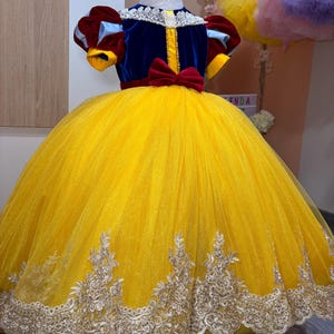 Peut inclure: Robe de princesse jaune en tulle avec un corsage en velours bleu, un nœud rouge et un col en dentelle blanche. La robe présente des détails brodés dorés à l'ourlet et des manches courtes rouges et bleues. Une tête de mannequin rose est au sommet.