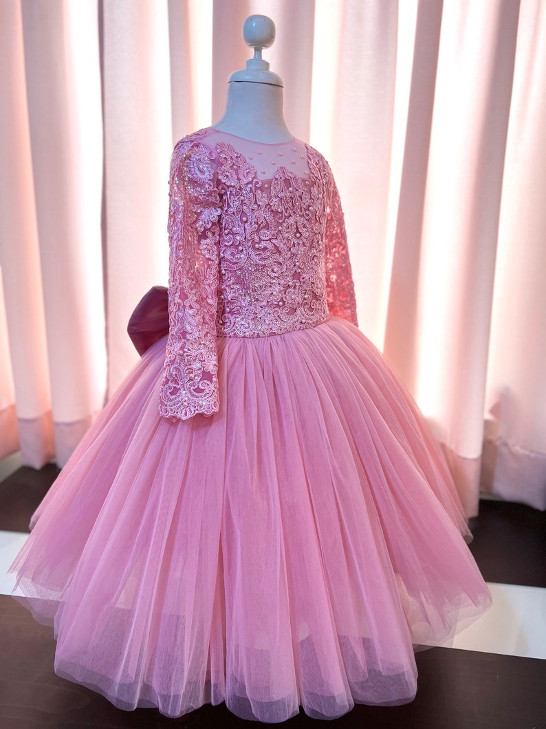 May include: A pink tulle dress with long sleeves and a lace bodice. The dress has a bow on the back and is perfect for a special occasion.