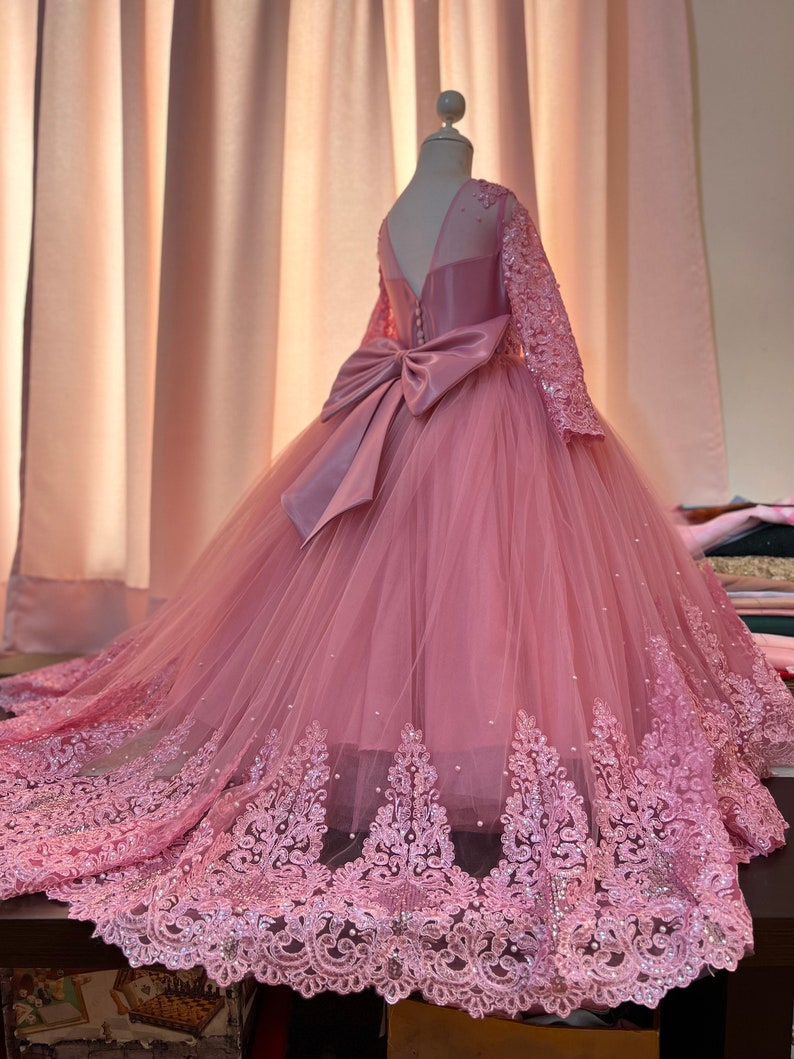 May include: A pink tulle dress with long sleeves and a lace trim. The dress has a satin bow at the back and is decorated with sequins.