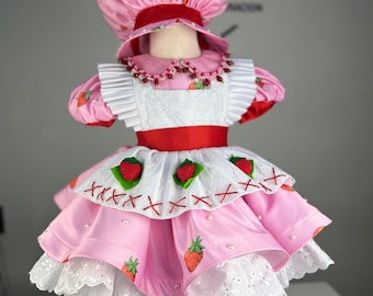 Op de afbeelding: Een roze en witte aardbeien-jurk met een bijpassende roze hoed. De jurk heeft een witte kantversiering en een rood lint om de taille. De hoed heeft een rood lint om de kroon.