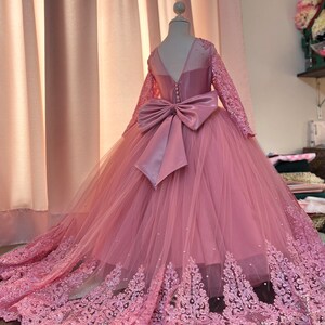May include: A pink tulle dress with long sleeves and a lace trim. The dress has a large satin bow at the back.