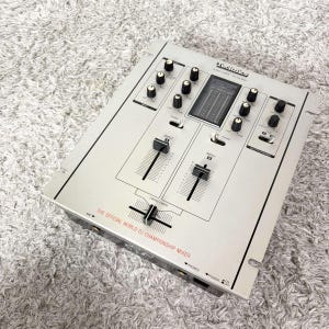 Peut inclure: Une table de mixage audio Technics SH-DX1200 argentée avec plusieurs boutons rotatifs noirs, deux faders de canal verticaux et un crossfader horizontal. La façade affiche le texte THE OFFICIAL WORLD DJ CHAMPIONSHIP MIXER en rouge.