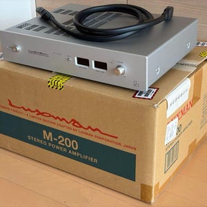 May include: A silver Luxman M-200 stereo power amplifier with a black power cable coiled on top. The amplifier features a front panel with two digital displays, a power switch, and a volume knob. It sits on a cardboard box labelled Luxman M-200.