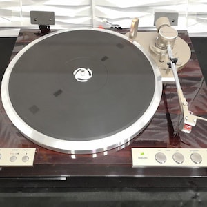 Może przedstawiać: Gramofon z ciemnego drewna z srebrnym talerzem i ramieniem. Gramofon ma srebrny panel sterowania z przyciskami do regulacji prędkości, powtarzania i innych funkcji.