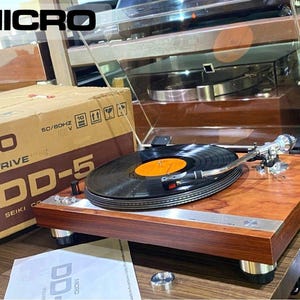 Pode incluir: Um gira-discos de tração direta Micro Seiki DD-5 com base em madeira, braço prateado e prato preto com um disco de vinil de etiqueta laranja. O gira-discos está posicionado ao lado da sua caixa de cartão original com texto impresso.