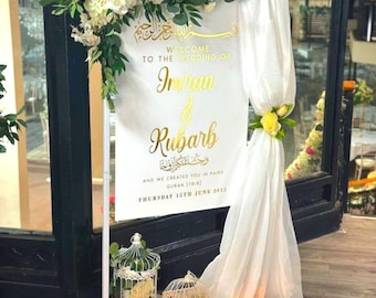 Può includere: Un cartello di benvenuto per matrimonio bianco con lettere dorate, con i nomi Imran & Rubarb, la data giovedì 15 giugno 2023, e il testo "Benvenuti al matrimonio di" e "E vi abbiamo creati in coppia Corano (78:18)". Il cartello è decorato con fiori bianchi e verde, ed è circondato da tessuto bianco e candele.