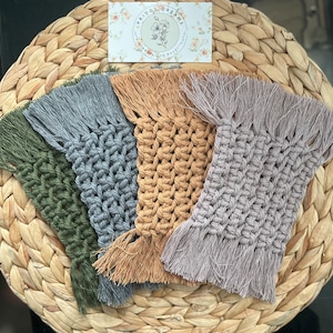 May include: Four crocheted coasters with fringe in various colors: green, gray, beige, and purple. The coasters are arranged in a semi-circle on a woven surface. The text "Priddy Dreams" is visible on a card in the background.