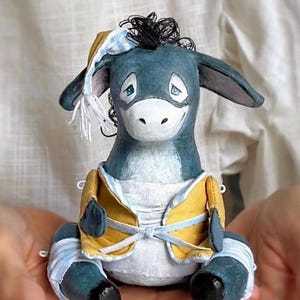 May include: A handmade blue donkey doll with a yellow vest and a striped hat. The doll has black hair and blue and white striped bandages on its legs. The donkey is sitting and has a smiling expression.