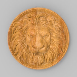 Op de afbeelding: Een ronde houten plaquette met een gegraveerd leeuwenhoofd in het midden. De leeuwenmanen zijn gedetailleerd en het gezicht is woest.