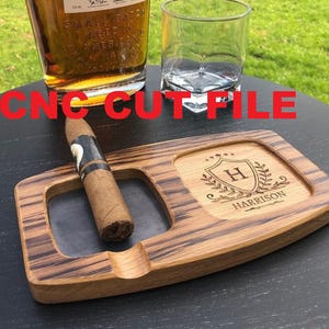 May include: A wooden cigar ashtray with a personalized engraving that reads "Harrison" and a monogram "H". A cigar rests in the ashtray. A glass of whiskey and an empty glass are also visible. The text "CNC CUT FILE" is in red.