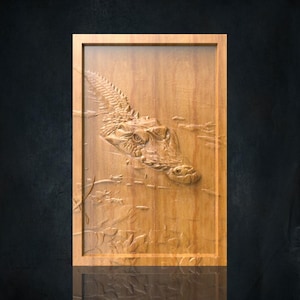 Op de afbeelding: Rechthoekig houten wandkunstwerk met een gedetailleerde reliëfsnede van een alligatorhoofd dat uit het water oprijst. Het kunstwerk is ingelijst en heeft een natuurlijke houtnerf, met details van de aquatische omgeving.